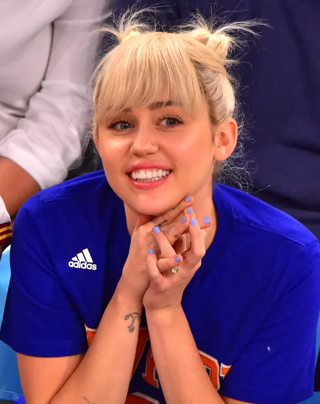 A person with blonde hair styled in buns, wearing a blue sports jersey, smiling with their hands resting under their chin.