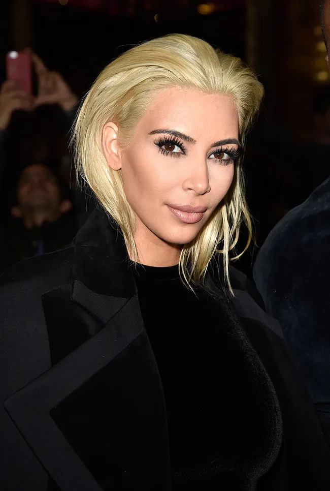 Blonde woman in a black outfit with smoky eye makeup, smiling slightly.