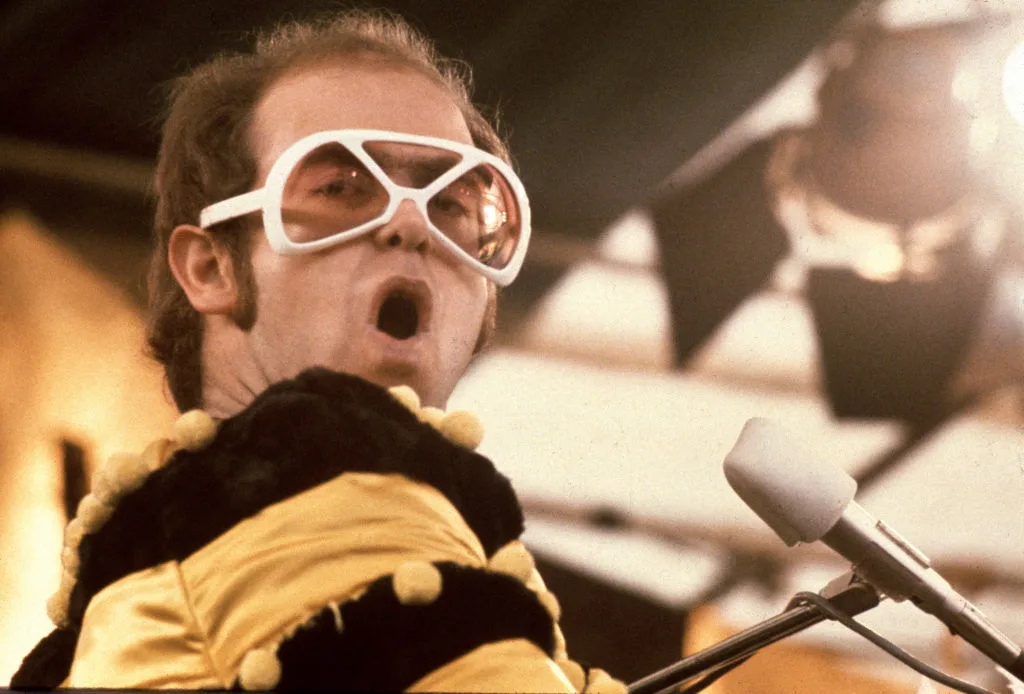 Elton John on stage with white glasses and a yellow and black jumper