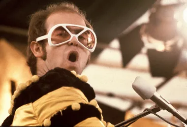 Elton John on stage with white glasses and a yellow and black jumper