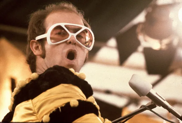 Elton John on stage with white glasses and a yellow and black jumper