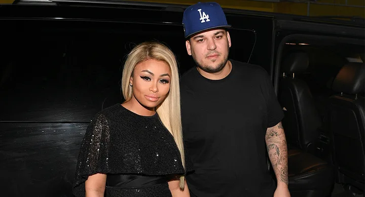 Two people standing in front of a vehicle; one wears a black shirt and cap, the other a sequined black outfit.