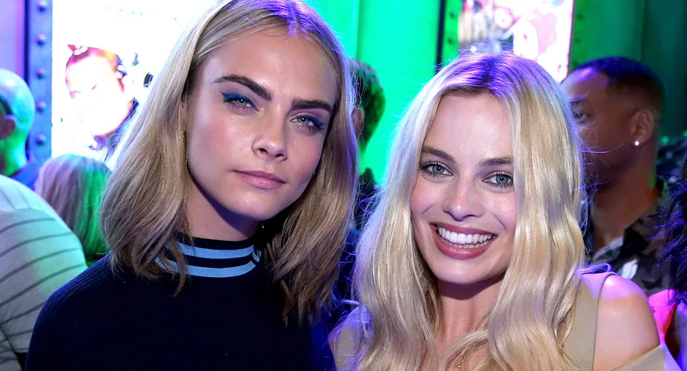 Two women with blonde hair smiling at a brightly lit event, with colorful backgrounds and people in the distance.