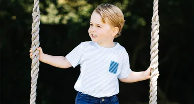 Young child with short, light hair, holding onto rope swings, wearing a striped shirt, smiling outdoors.