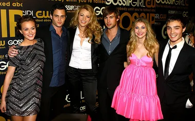 Cast members of "Gossip Girl" posing at an event, standing in front of a branded backdrop, smiling and dressed up.