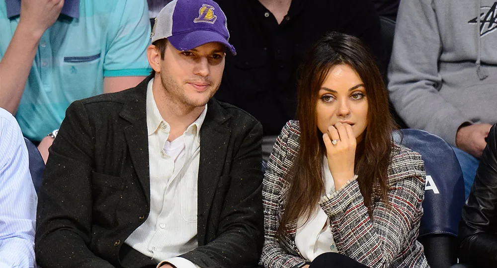 A man in a Lakers cap and a woman sit courtside at a basketball game, surrounded by other spectators.