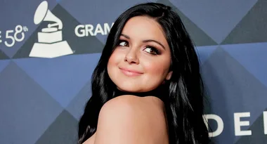 A woman with long dark hair smiles over her shoulder at an event with a Grammy Award backdrop.