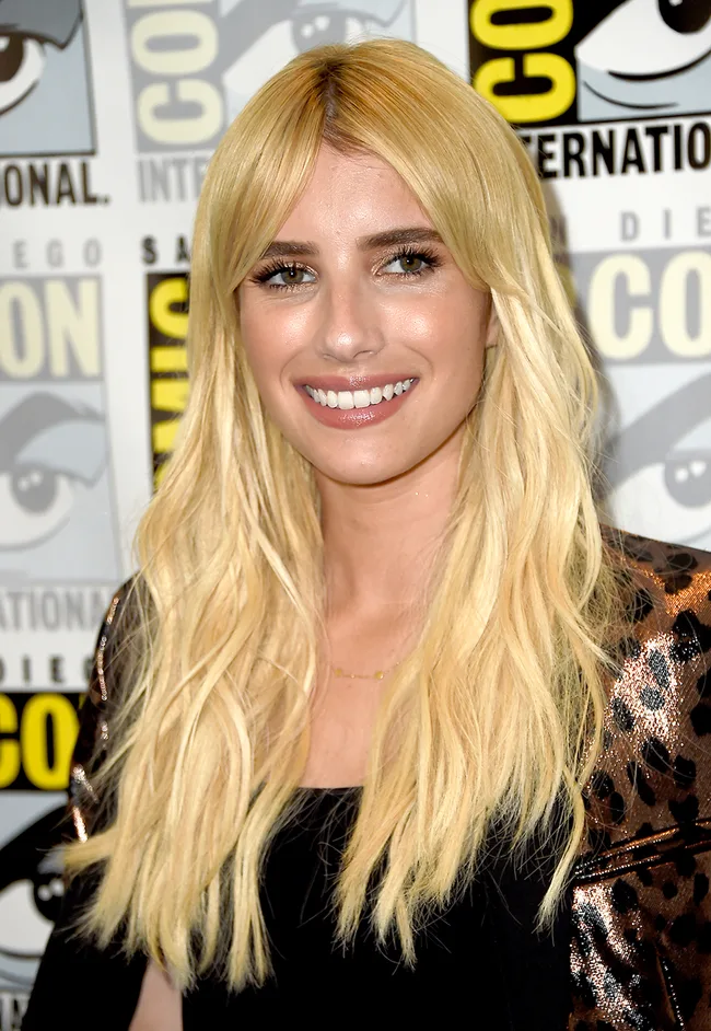 Blonde woman smiling at Comic-Con event with logo backdrop, wearing a black top and patterned jacket.