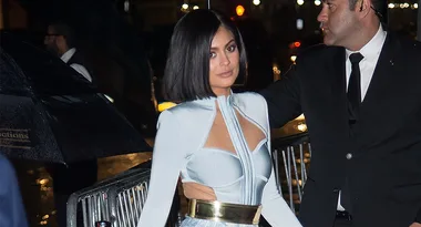 A woman in a light blue dress with cutouts, escorted by a man in a suit, outside at night.