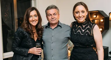 A man and two women smiling, standing indoors, with one woman holding a glass.