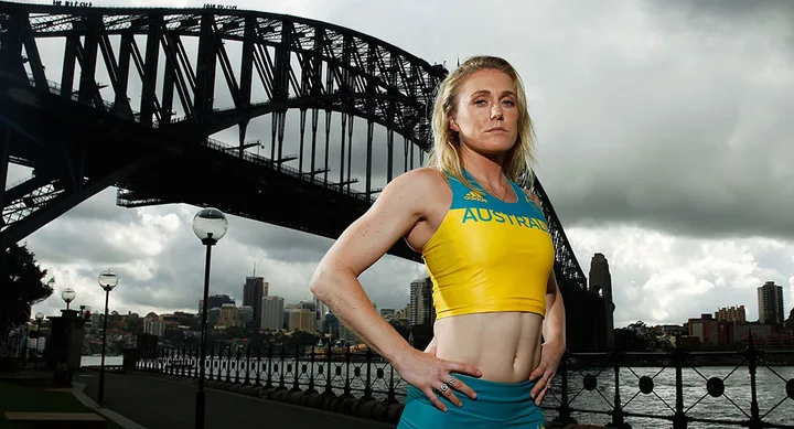 Female athlete in Australian sportswear stands confidently, with hands on hips, in front of Sydney's Harbour Bridge.
