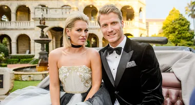 Two elegantly dressed individuals seated in a vintage car outside a grand building with a fountain in the background.