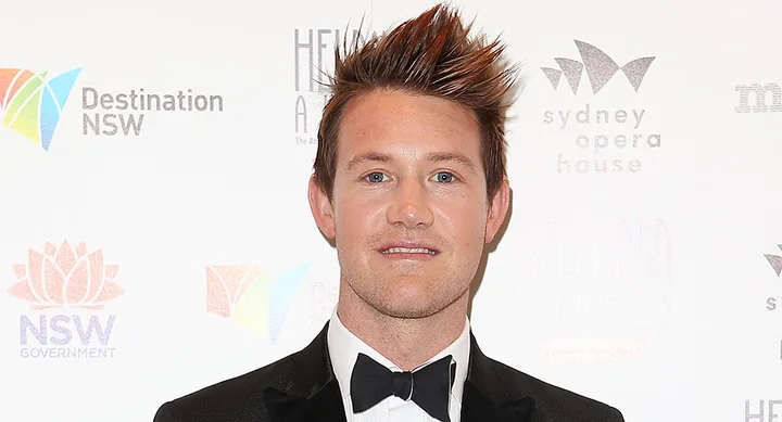 Man in a tuxedo with spiked hair at an event with a backdrop featuring logos of NSW and Sydney Opera House.