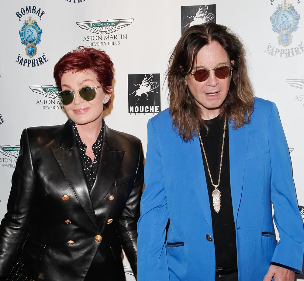 Two people wearing sunglasses at a branded event; one in a black leather jacket, the other in a blue blazer.