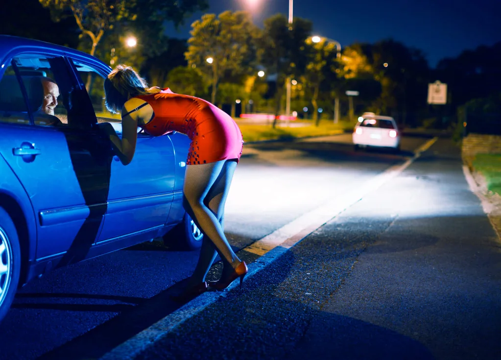 A sex worker talking to a man through a blue car's window