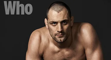 Shirtless man with a serious expression poses against a dark background with the word "Who" in the corner.