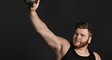 Man in black tank top lifting a kettlebell against a dark background.
