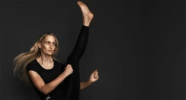 A woman performing a high karate kick in a black outfit against a dark background.