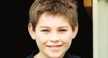 A young child with short brown hair and a slight smile, standing against a dark background.