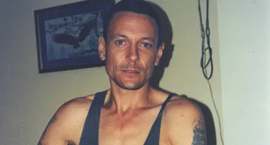 Man in a tank top poses indoors with a framed picture in the background.
