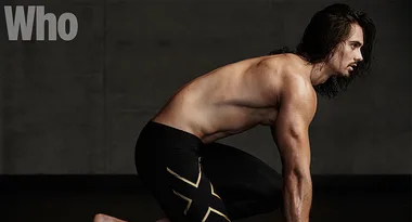A shirtless man in athletic posture on a dark background, wearing black compression shorts with gold accents.