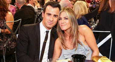 A man in a suit and a woman in a strapless dress smiling, sitting together at an event with people in the background.