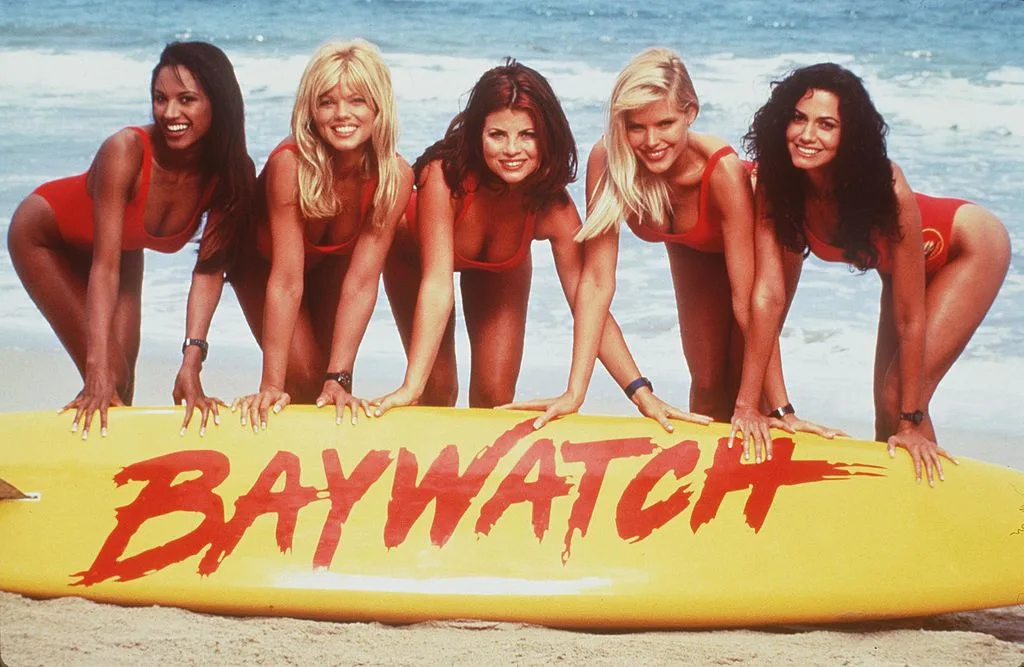 Girls of Baywatch posing in fronnt of a surfboard with the writing 'Baywatch'