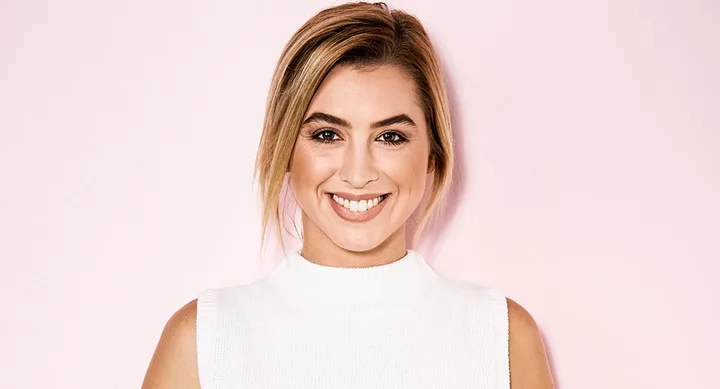 A woman with light brown hair smiles at the camera against a pale pink background wearing a sleeveless white top.