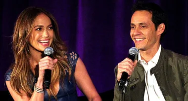 Two people smiling and holding microphones during a talk, seated in front of a dark purple curtain.