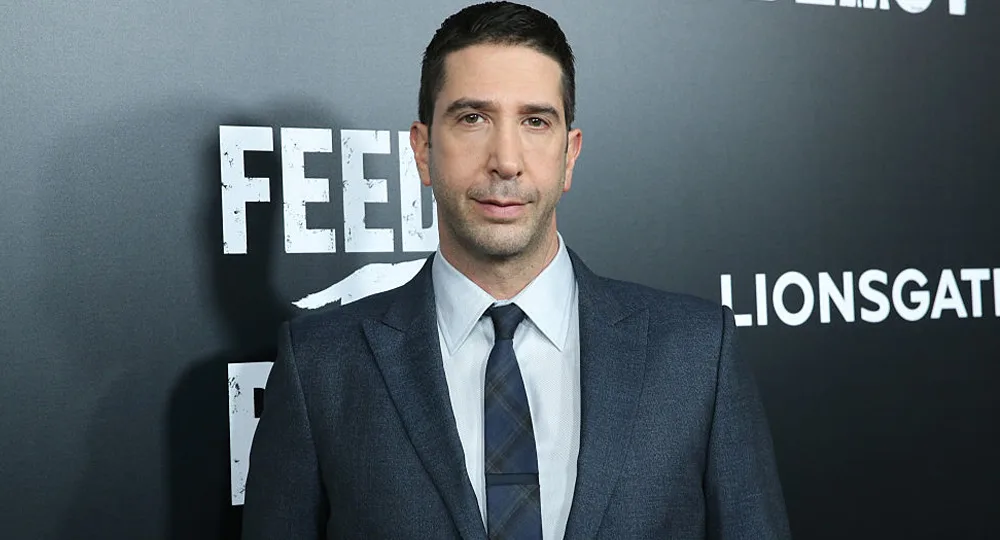 A man in a suit stands in front of a black backdrop with white text at a movie premiere or event.