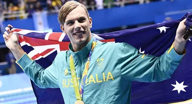 Australian athlete celebrates with flag at an Olympic event, wearing a gold medal and green jacket with "Australia" text.