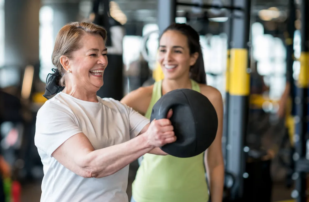A mature woman lifting a kettle-ball next to her personal trainer