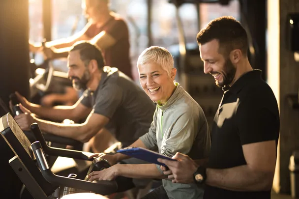 A male personal trainer standing next to a mature woman on a treadmill