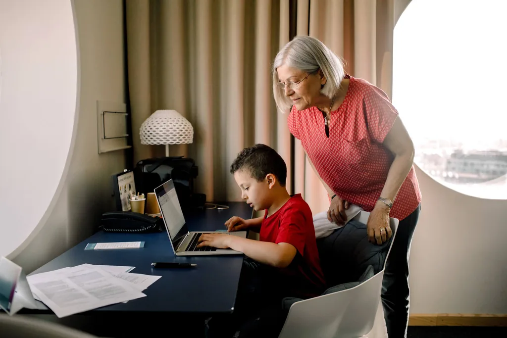 A grandmother looking over a young boy's shoulder while he plays on his laptop