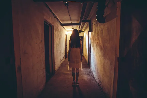 A person in a white dress stands in a dimly lit, narrow corridor with concrete walls.