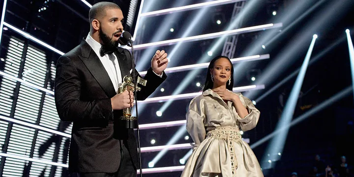 Man in a tuxedo speaks into a microphone holding a trophy, alongside a woman in an elegant dress, on a stage with bright lights.