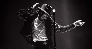 Singer on stage in a fedora and sequined jacket, performing a dance move with a spotlight highlighting his pose.