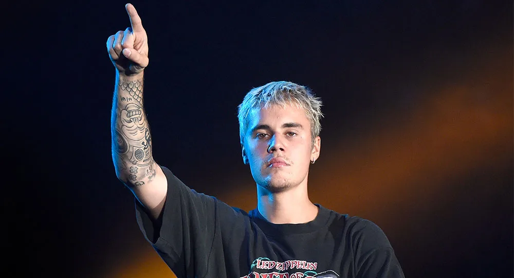 Singer with tattoos points upward, wearing a Led Zeppelin T-shirt, on stage with a spotlight backdrop.