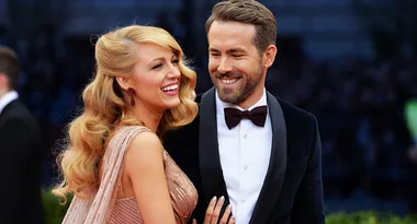 A couple smiling and posing on a red carpet, with the woman in a pink gown and the man in a tuxedo and bow tie.