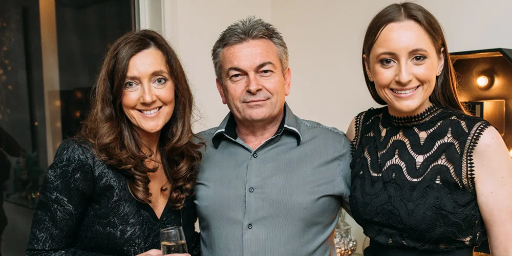 Three people smiling at a gathering, two women in black dresses and a man in a gray shirt, holding drinks.