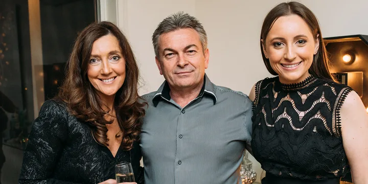 Three people smiling at a gathering, two women in black dresses and a man in a gray shirt, holding drinks.