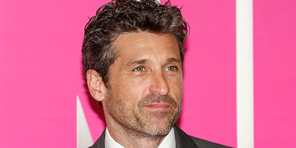 A person in a suit stands before a bright pink background, looking to their left.