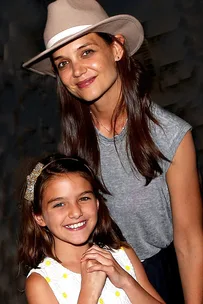 Two people smiling; one in a hat and gray shirt, the other in a white dress with a gold bow headband.