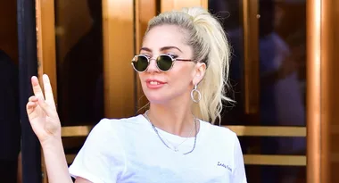 A person with sunglasses gives a peace sign, wearing a white t-shirt and hoop earrings, in front of a building.