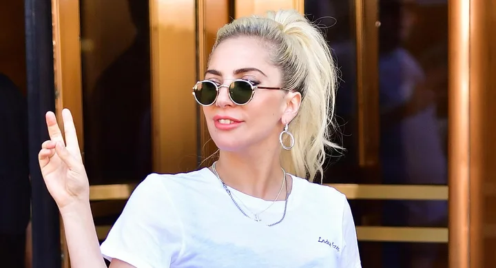 A person with sunglasses gives a peace sign, wearing a white t-shirt and hoop earrings, in front of a building.