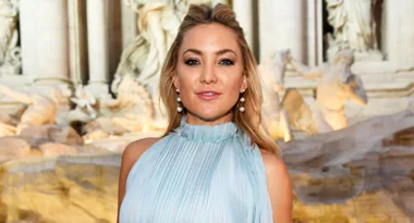 A woman in a light blue dress poses in front of a grand stone fountain with sculptures.