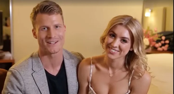 A smiling couple seated indoors, with soft lighting and a bouquet in the background.