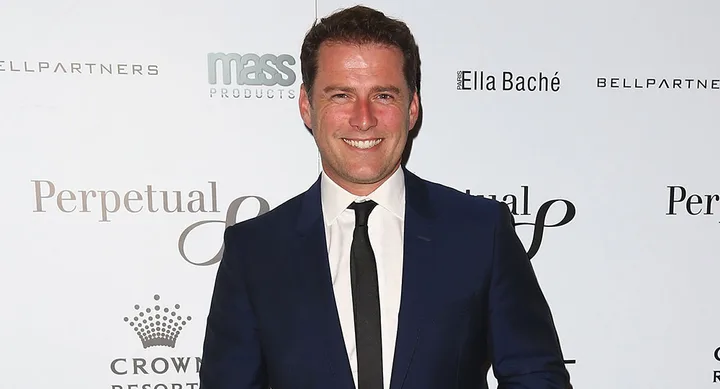 Man in a dark suit and tie smiling at an event with branded backdrop.