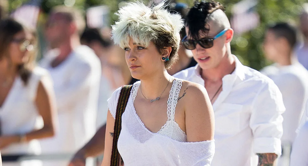 Two individuals in white outfits walking outdoors; one with short blond hair, the other with a mohawk and sunglasses.
