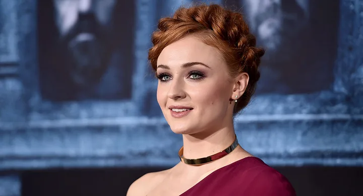 A woman with braided red hair and a metallic collar poses on a dark, patterned background at an event.